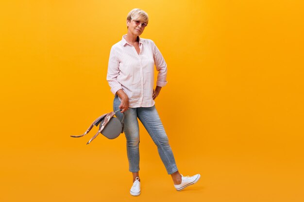 Kurzhaarige Dame in Jeans und Hemd wirft mit Handtasche auf orange Hintergrund auf