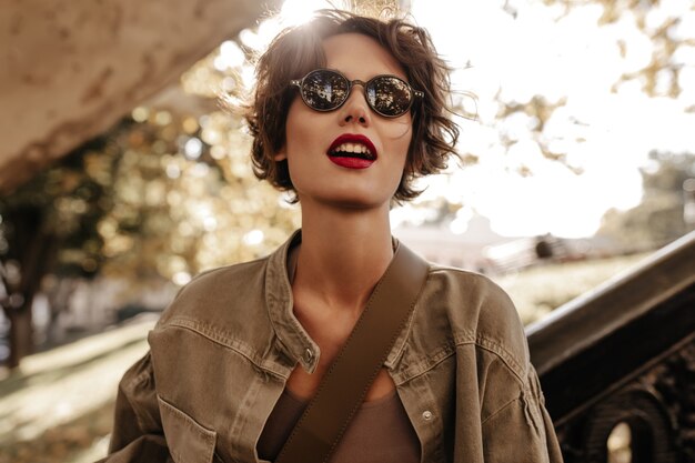 Kurzhaarige Frau mit schwarzer Sonnenbrille in olivgrüner Jacke schaut nach draußen. Wunderbare Frau mit hellem Lippenstift, der draußen aufwirft.