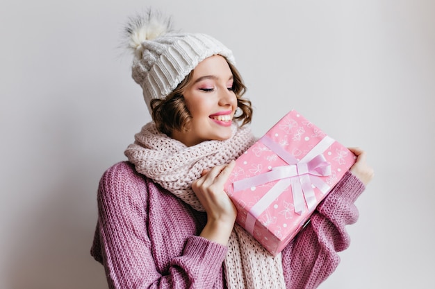 Kurzhaariges europäisches Mädchen im Hut, das emotional aufwirft und Neujahrsgeschenk betrachtet. Innenfoto der verblüfften jungen Dame im niedlichen Schal, der rosa Geschenk hält.