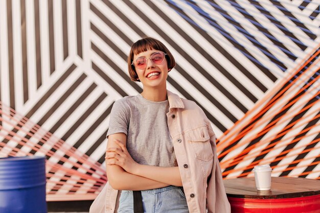 Kurzhaariges Mädchen mit rosa Sonnenbrille lächelnd im gestreiften HintergrundTeenager mit brünetten Haaren in grauem T-Shirt und beiger Jacke posiert im Café