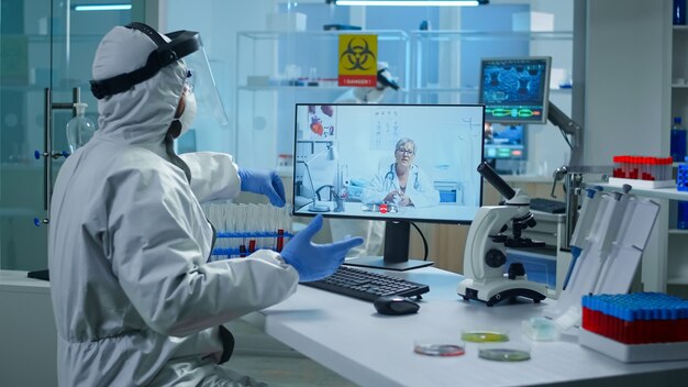Labortechniker im PPE-Anzug, der am Tisch im medizinischen Forschungslabor sitzt, ein Reagenzglas hält und per Videoanruf mit dem leitenden Arzt spricht, der während des virtuellen Treffens Online-Ratschläge gibt