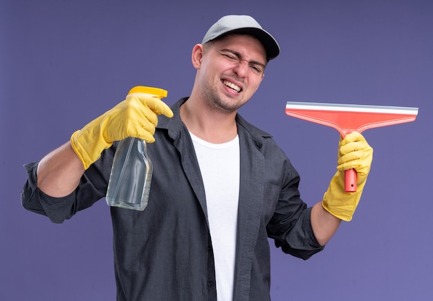 Lachen mit geschlossenen Augen junger hübscher Reinigungsmann, der T-Shirt mit Kappe und Handschuhen hält, die Moppkopf und Sprühflasche lokalisiert auf lila Wand halten