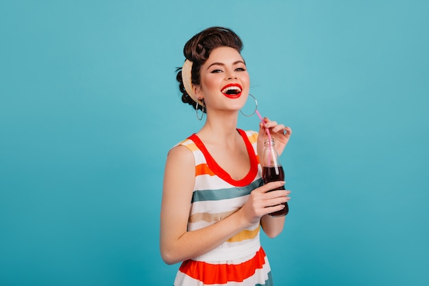 Lachende Frau im gestreiften Kleid, die Soda trinkt. Studioaufnahme des glücklichen Pinup-Mädchens mit Getränk lokalisiert auf blauem Hintergrund.