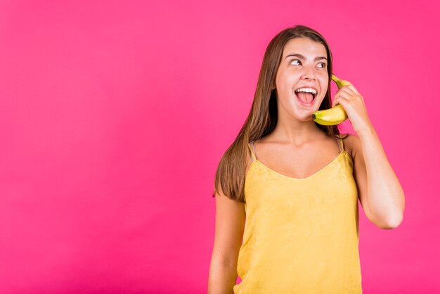 Lachende junge Frau, die auf Banane auf rosa Hintergrund spricht