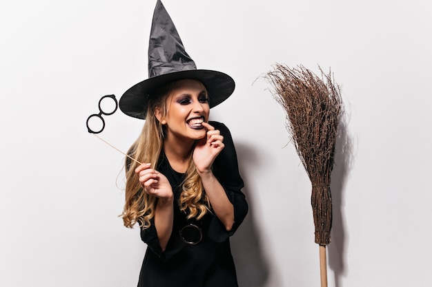 Lachende lockige Hexe, die Halloween genießt. Innenporträt des gut gelaunten Zauberers lokalisiert auf weißer Wand.