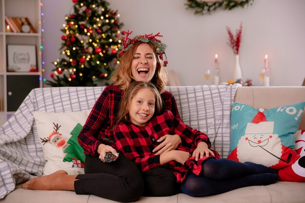 Lachende Mutter mit Stechpalmenkranz hält TV-Fernbedienung und schaut in die Kamera mit Tochter, die auf der Couch sitzt und die Weihnachtszeit zu Hause genießt