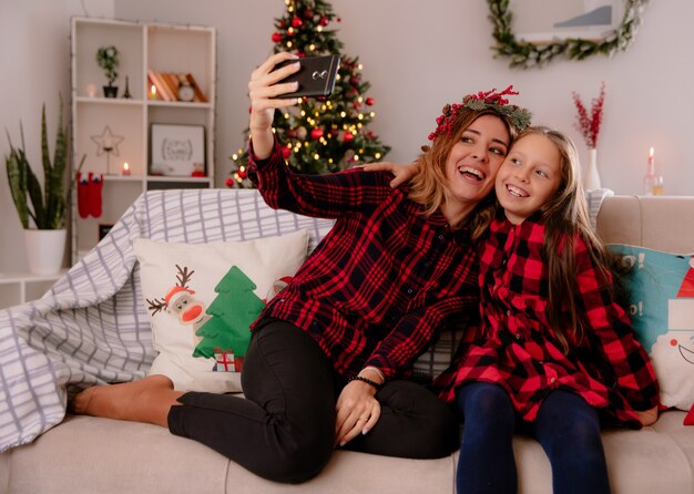 Lachende Mutter mit Stechpalmenkranz und Tochter schauen auf Telefon, das Selfie nimmt, das auf Couch sitzt und Weihnachtszeit zu Hause genießt