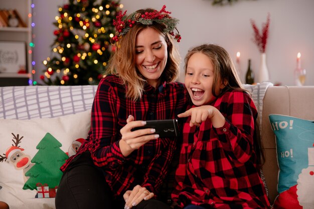 Lachende Mutter und Tochter beobachten etwas am Telefon sitzen auf der Couch und genießen die Weihnachtszeit zu Hause