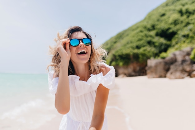Lachende spektakuläre Frau in der Sonnenbrille, die Urlaub auf tropischer Insel genießt. Foto im Freien der liebenswerten Frau im weißen Kleid, das auf Natur lächelt.