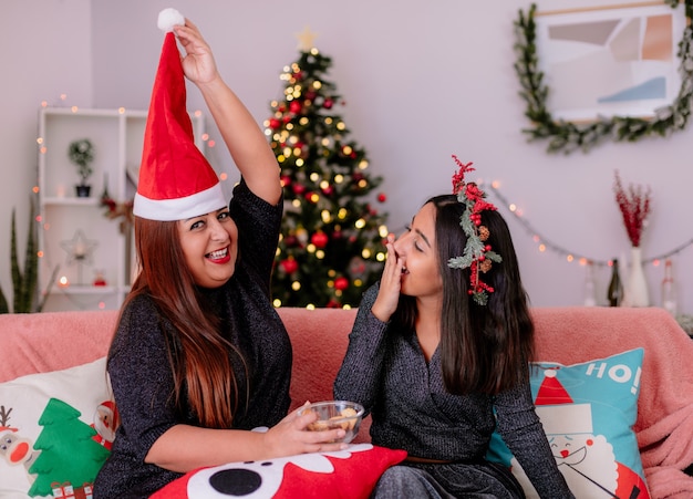 Lachende Tochter mit Stechpalmenkranz schaut auf die Mutter, die eine Weihnachtsmütze über dem Kopf hält und auf der Couch sitzt und die Weihnachtszeit zu Hause genießt