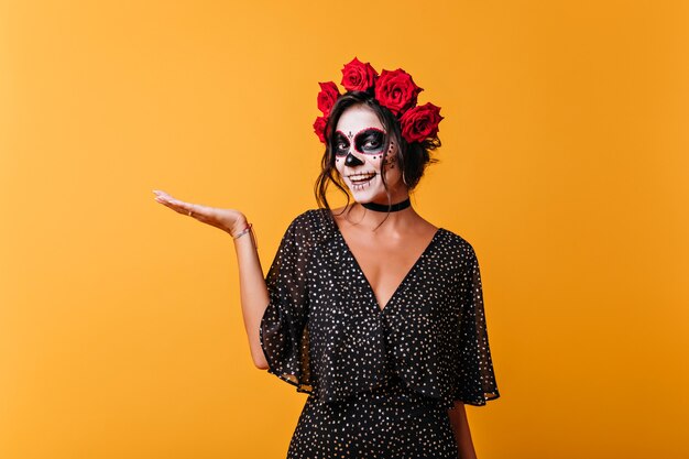 Lachende Zombiedame, die auf gelbem Hintergrund aufwirft. Atemberaubendes weibliches Modell in der mexikanischen Halloween-Kleidung, die zur Kamera lächelt.