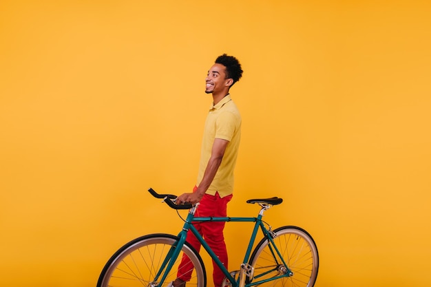 Lachender afrikanischer junger Mann in stylischen Hosen posiert mit Fahrrad Studioaufnahme eines gutaussehenden männlichen Models, das mit einem Lächeln auf gelbem Hintergrund steht