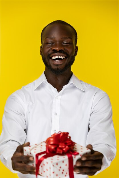 Lachender bärtiger junger afroamerikanischer Kerl hält ein Geschenk in zwei Händen im weißen Hemd