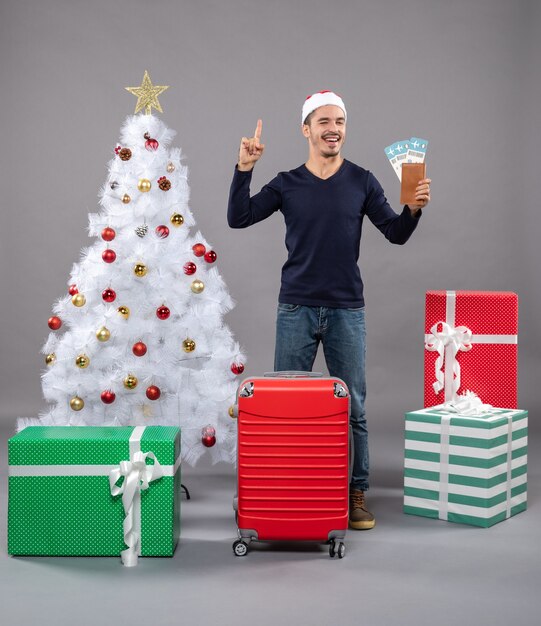 lachender Mann mit rotem Koffer, der seine Reisetickets hält und etwas auf Grau zeigt