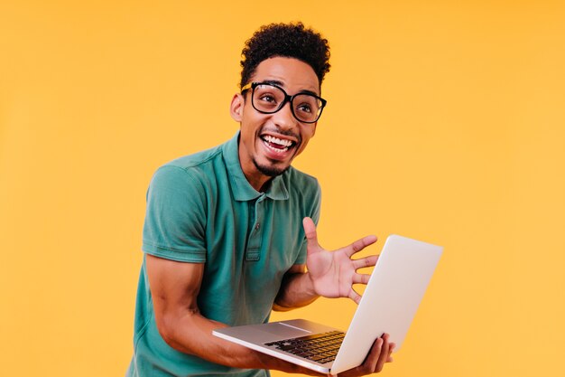 Lachender schwarzer Mann in Gläsern, die Aufregung ausdrücken. emotionaler internationaler Student, der Computer hält.
