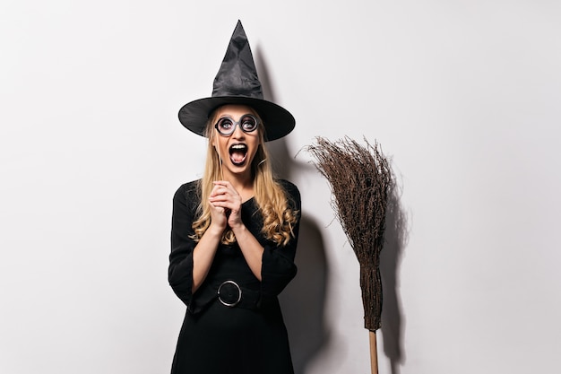 Kostenloses Foto lachendes blondes mädchen, das maskerade in halloween genießt. gut gelaunte hexe posiert im schwarzen hut.