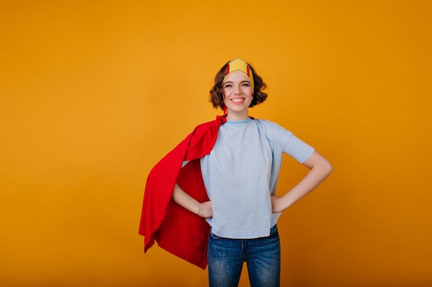 Lachendes brünettes Mädchen im Superhelden-Outfit, das auf gelbem Raum aufwirft