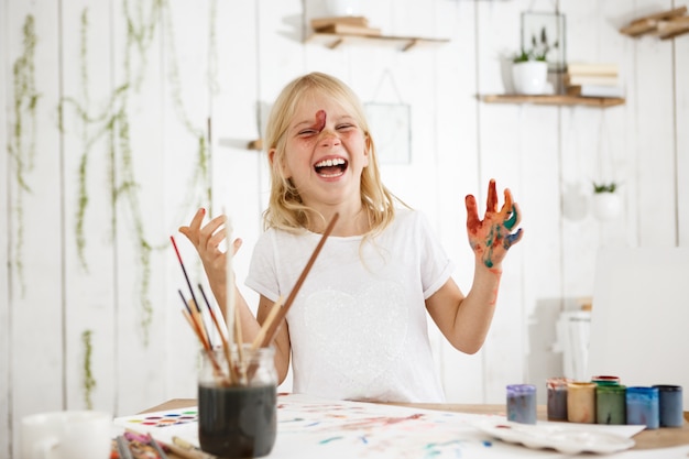 Lachendes, charmantes kleines Mädchen mit blonden Haaren, Sommersprossen und blauen Augen hat sich mit Farbe durcheinander gebracht. Kreatives Kind mit Farbe auf Gesicht und Händen.