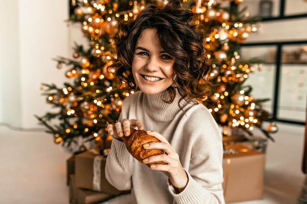 Lachendes glückliches Mädchen mit lockiger Frisur, die mit Croissant über Weihnachtsbaum mit glücklichen Gefühlen aufwirft. Neujahrsmorgen, Weihnachtsfeier