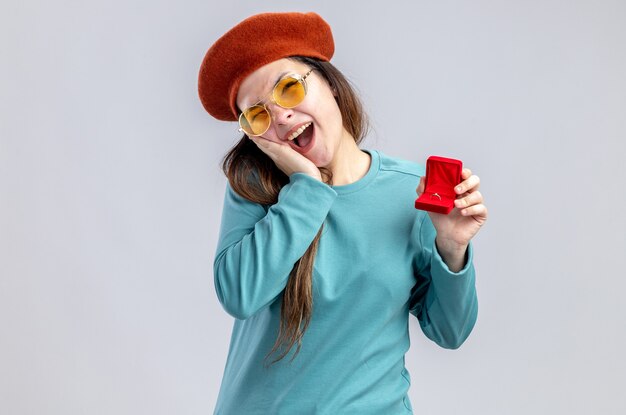 Lachendes junges Mädchen am Valentinstag mit Hut mit Brille mit Ehering, der Hand auf die Wange legt, isoliert auf weißem Hintergrund