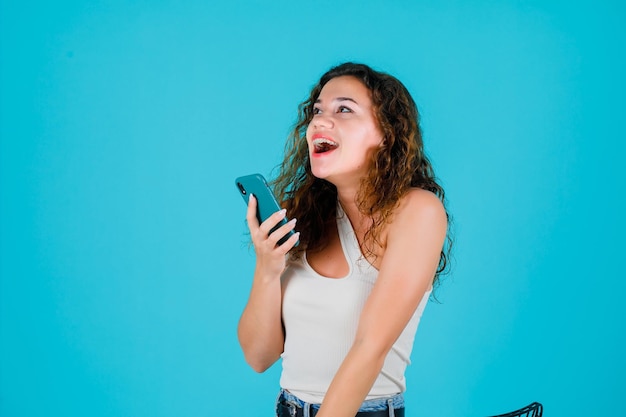 Lachendes Mädchen schaut nach oben, indem es das Telefon auf blauem Hintergrund hält
