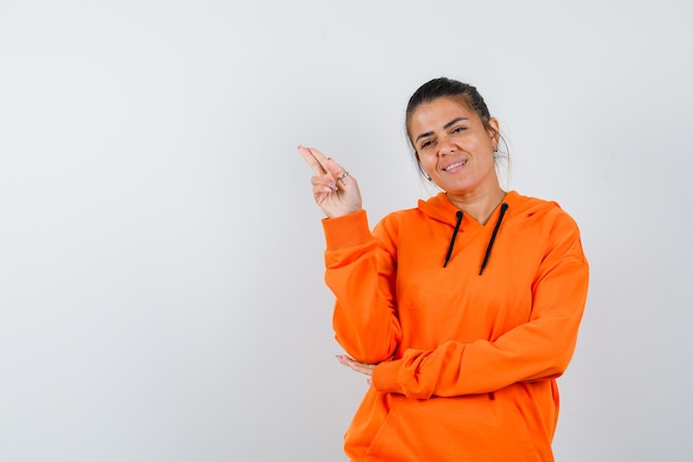 Lady gestikuliert mit hand und zwei fingern im orangefarbenen hoodie und sieht zufrieden aus