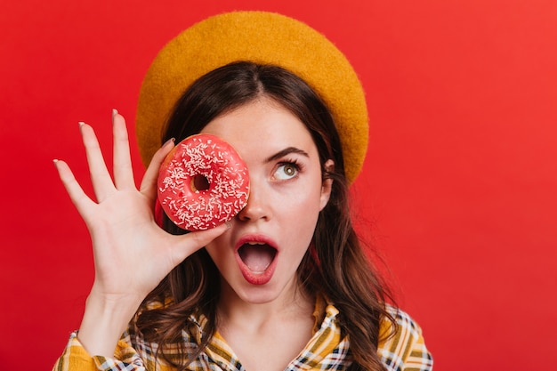 Kostenloses Foto lady schaut überrascht auf und bedeckt ihre augen mit appetitlichem donut. mädchen im orangefarbenen hut im staunen, das auf roter wand aufwirft.