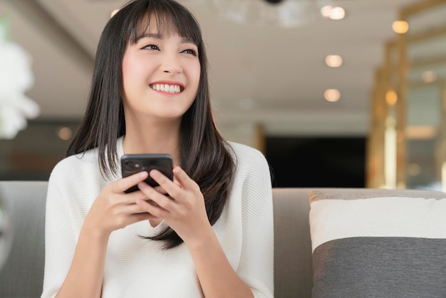 Lächeln Glück fröhliche asiatische weibliche Hand verwenden Smartphone denken und schauen mit freudiger Smartphone-Technologie und Kommunikationsideen-Konzept nach Hause Hintergrund