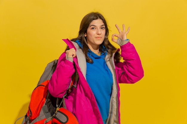 Lächeln, zeigt sich schön. Porträt eines fröhlichen jungen kaukasischen Touristenmädchens mit Tasche und Fernglas lokalisiert auf gelbem Studiohintergrund.