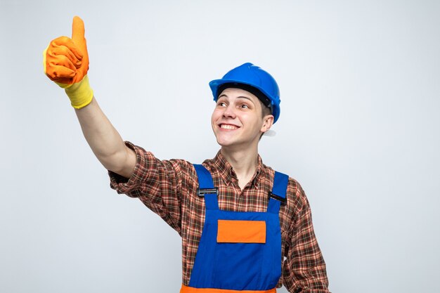 Lächelnd aussehende Seite mit Daumen nach oben junger männlicher Baumeister, der Uniform mit Handschuhen trägt