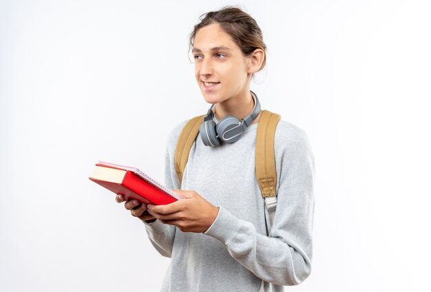 Lächelnd aussehender junger Kerl Student mit Rucksack mit Kopfhörern am Hals, der Bücher hält?