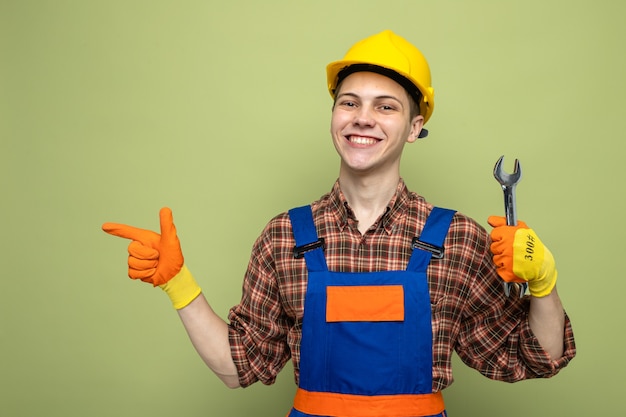 Lächelnd aussehender junger männlicher Baumeister, der Uniform mit Handschuhen trägt, die einen Gabelschlüssel halten