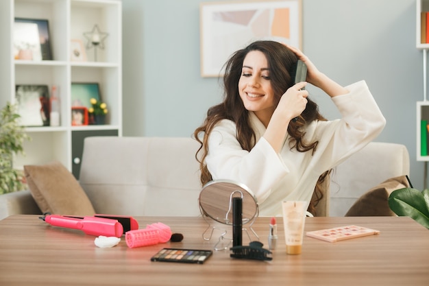 Lächelnd aussehender Spiegel junges Mädchen, das Haare kämmt, die am Tisch mit Make-up-Tools im Wohnzimmer sitzen?