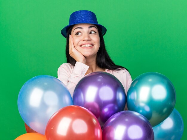 Lächelnd aussehendes junges schönes Mädchen mit Partyhut, das hinter Ballons steht und die Hand auf die Wange legt, isoliert auf grüner Wand