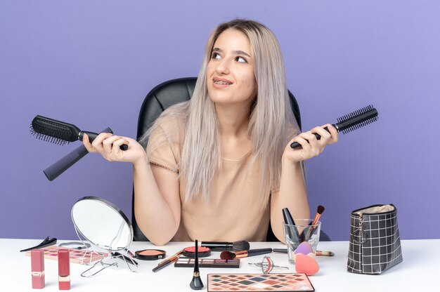 Lächelnd aussehendes junges schönes Mädchen sitzt am Tisch mit Make-up-Tools, die den Kamm einzeln auf blauem Hintergrund halten