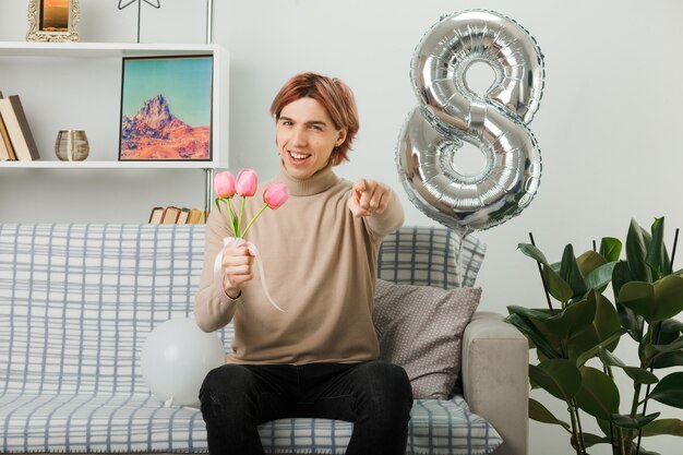 Lächelnd, das Ihnen zeigt, dass Sie am glücklichen Frauentag einen hübschen Kerl gestikulieren, der Blumen auf dem Sofa im Wohnzimmer hält?