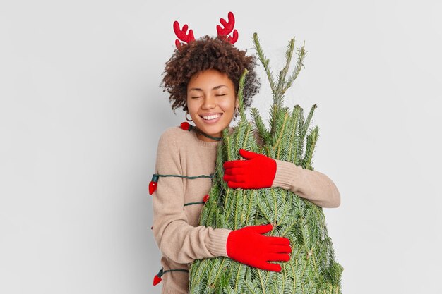Lächelnd entzückte afroamerikanische Frau trägt Rentierhörner und Handschuhe umarmt grünen Tannenbaum mit Liebe, die glücklich ist, Neujahr zu Hause zu feiern, kehrt vom Weihnachtsstraßenmarkt zurück, der von Girlande umwickelt wird