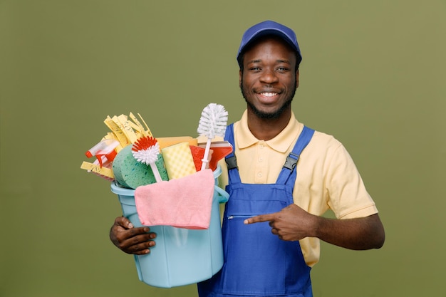 Lächelnd halten und zeigt auf Eimer mit Reinigungswerkzeugen junger afroamerikanischer Reinigungsmann in Uniform mit Handschuhen isoliert auf grünem Hintergrund