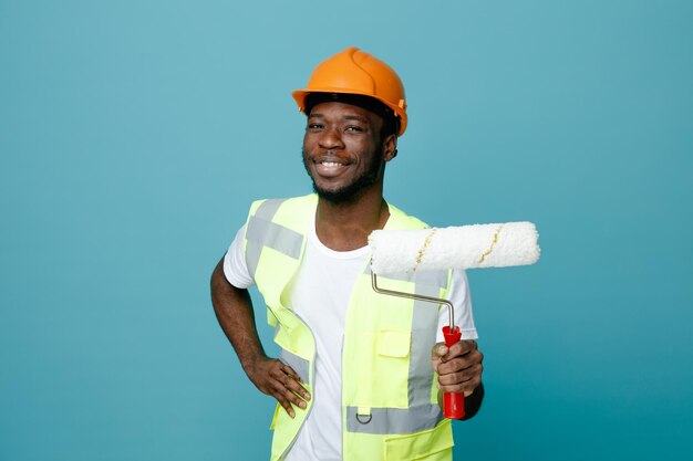Lächelnd, Hand auf die Hüften legend, junger afrikanisch-amerikanischer Baumeister in Uniform, der Walzenbürste isoliert auf blauem Hintergrund hält