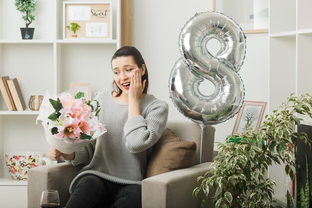 Lächelnd Hand auf Wange legen schönes Mädchen am glücklichen Frauentag mit Blumenstrauß auf Sessel im Wohnzimmer sitzend