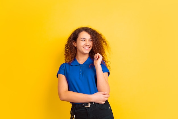 Lächelnd, lachend. Mädchenporträt des kaukasischen Teenagers auf gelbem Studiohintergrund. Schönes weibliches lockiges Modell im blauen Hemd. Konzept der menschlichen Emotionen, Gesichtsausdruck, Verkauf, Anzeige. Exemplar.