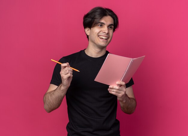 Lächelnd mit Blick auf die Seite junger gutaussehender Kerl mit schwarzem T-Shirt, das Notizbuch mit Bleistift hält, isoliert auf rosa Wand
