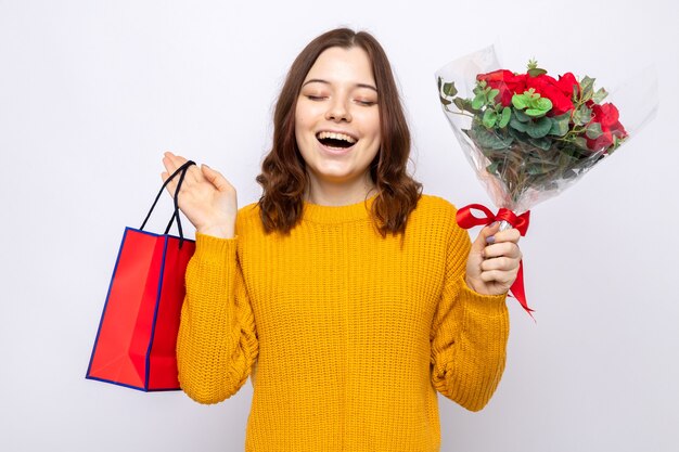 Lächelnd mit geschlossenen Augen schönes junges Mädchen am glücklichen Frauentag mit Geschenktüte mit Blumenstrauß isoliert auf weißer Wand