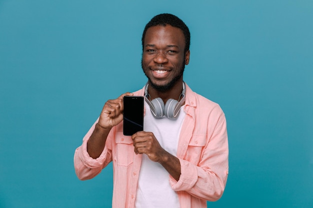 Lächelnd mit Telefon junger Afroamerikaner, der Kopfhörer am Hals trägt, isoliert auf blauem Hintergrund
