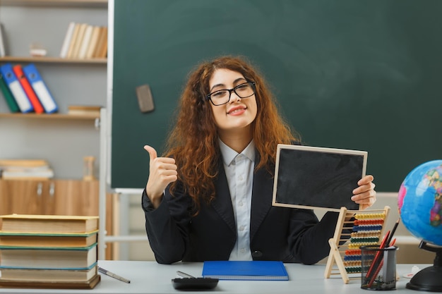 lächelnd zeigt Daumen nach oben Junge Lehrerin mit Brille und Mini-Tafel am Schreibtisch sitzend mit Schulwerkzeugen im Klassenzimmer