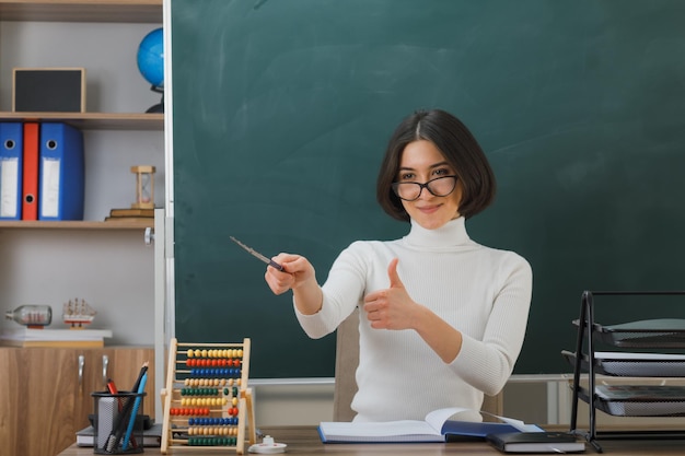 lächelnd zeigt Daumen nach oben Junge Lehrerin mit Brille zeigt an der Seite mit einem Zeiger, der am Schreibtisch mit Schulwerkzeugen im Klassenzimmer sitzt