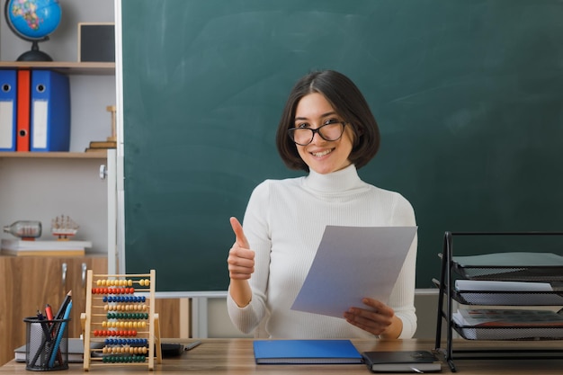 Lächelnd zeigt Daumen nach unten Junge Lehrerin mit Brille, die Papier hält und am Schreibtisch mit Schulwerkzeugen im Klassenzimmer sitzt