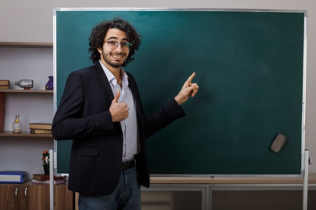 Lächelnd zeigt der junge männliche Lehrer mit Brille auf die Tafel im Klassenzimmer