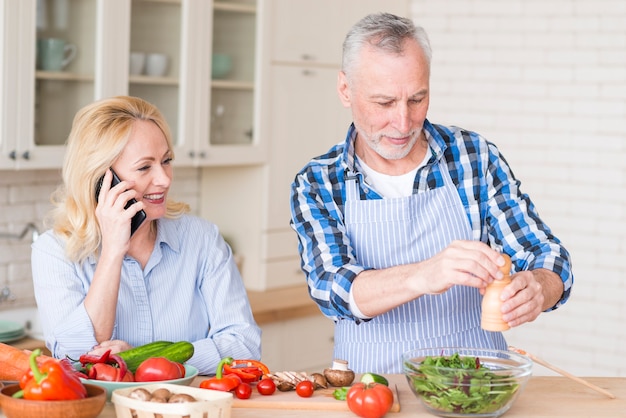 Lächelnde ältere Frau, die am Handy und ihrem Ehemann zubereitet den Salat in der Küche spricht