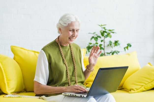 Lächelnde ältere Frau, die ihre Hand unter Verwendung des Laptops wellenartig bewegt